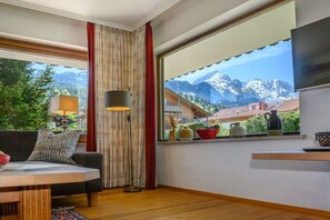 A room with a wooden floor, a sofa, a table, a lamp, a window with a view of mountains, and a curtain. - Holiday apartment with 2 bedrooms (Garmisch-Partenkirchen)