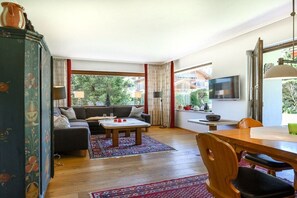 A living room with a sofa, coffee table, and dining table. There is a TV mounted on the wall and a window with curtains. - Holiday apartment with 2 bedrooms (Garmisch-Partenkirchen)