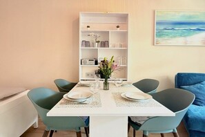 A dining area with a white table set for four, surrounded by teal chairs, a white bookshelf, and a painting of a beach scene. - Holiday apartment with 1 bedroom (Kressbronn am Bodensee)