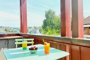 A balcony with a table, two drinks, a bowl of fruit, and a view of houses and trees. - Holiday apartment with 1 bedroom (Kressbronn am Bodensee)