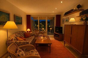A cozy living room with a sofa, a coffee table, and a television. There is a window with curtains and a view of the outdoors. - Garden apartment 283 (Lübeck-Travemünde)
