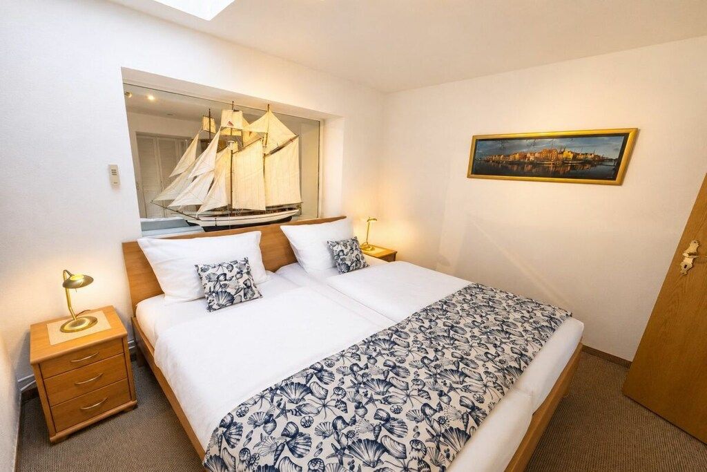 Une chambre d’hôtel avec un lit, une table de chevet, une lampe, une fenêtre qui reflète un voilier, et un tableau représentant un paysage urbain accroché au mur.