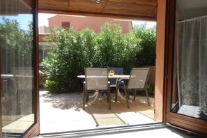 A patio area with a table and chairs, a small table with a bowl, and a view of a building and greenery outside. - Holiday home with 1 bedroom (Gruissan les Ayguades)
