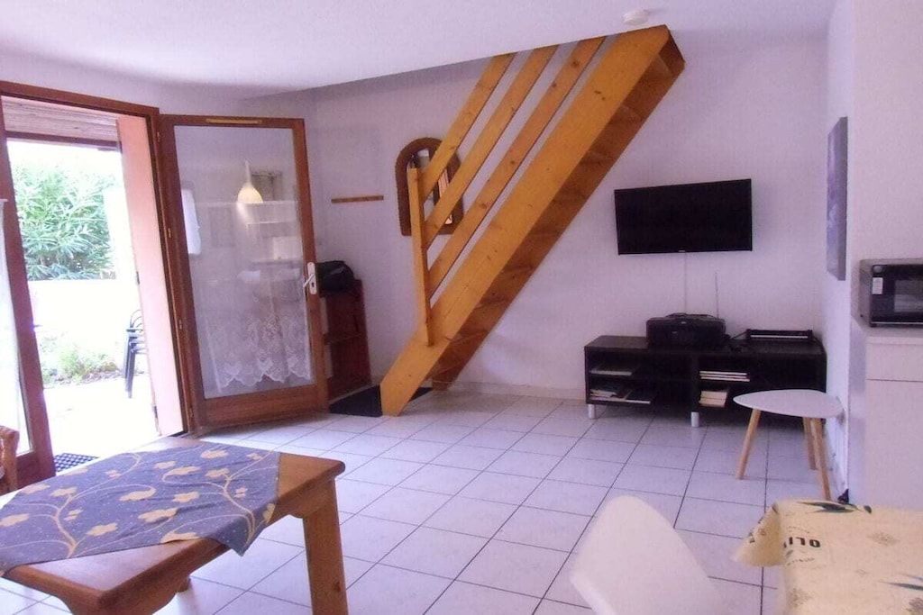 Une pièce avec un escalier en bois, un téléviseur fixé au mur, une table recouverte d’une nappe et une chaise.