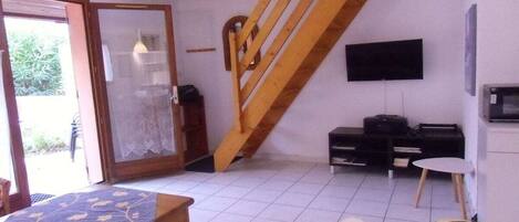 A room with a wooden staircase, a television mounted on the wall, a table with a tablecloth, and a chair.