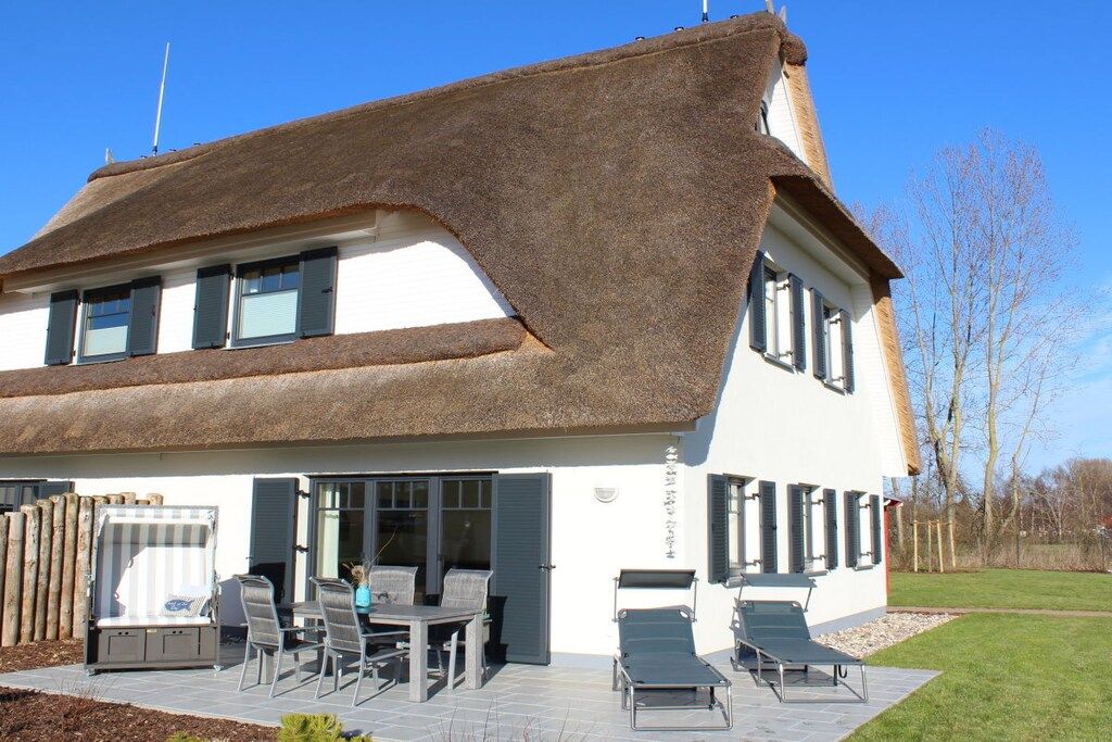 Uma casa de dois andares com telhado de palha, um pátio com móveis de exterior e um gramado bem cuidado.