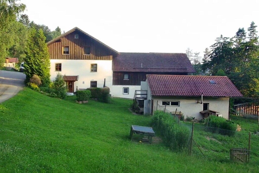 Une maison à deux étages avec un toit recouvert de tuiles rouges, un petit abri attenant et une cour gazonnée.