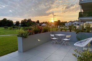 An outdoor patio with a table and chairs, a white chair, and a view of the sunset over a residential area. - Holiday apartment with 1 bedroom (Friedrichshafen)
