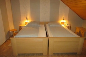 Habitación doble con dos camas individuales, cabeceros de madera, mesitas de noche con lámparas y techo inclinado.