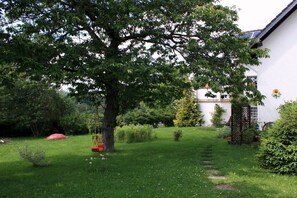 A well-maintained garden with a variety of trees and shrubs, a swing set, and a white building with a visible window and door. - Holiday apartment Sunflower with sauna and garden (Daun)