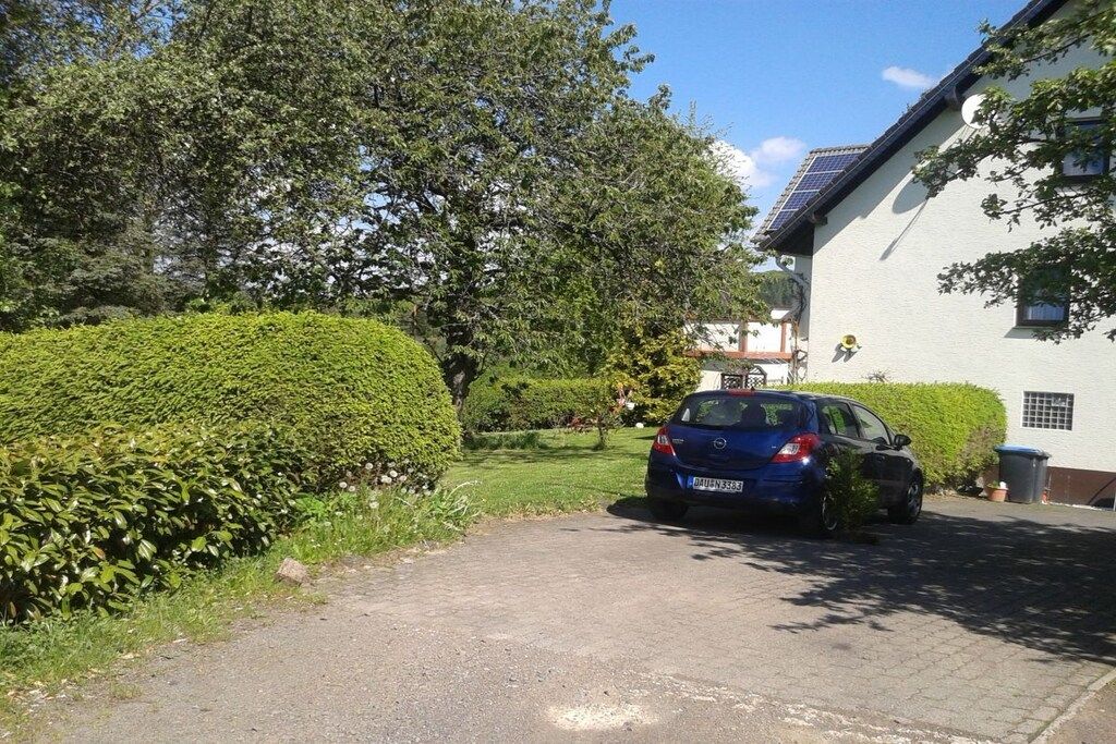 Un camino de acceso residencial con un coche estacionado, una casa blanca y un seto bien cuidado.