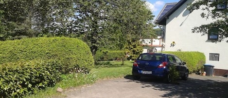 Un camino de acceso residencial con un coche estacionado, una casa blanca y un seto bien cuidado.