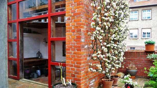 Een bakstenen muur met een bloeiende plant in een pot en een raam met rode omlijsting.
