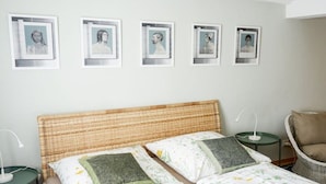 Een slaapkamer met een bed, kussens, een stoel, een nachtkastje en ingelijste foto's aan de muur.