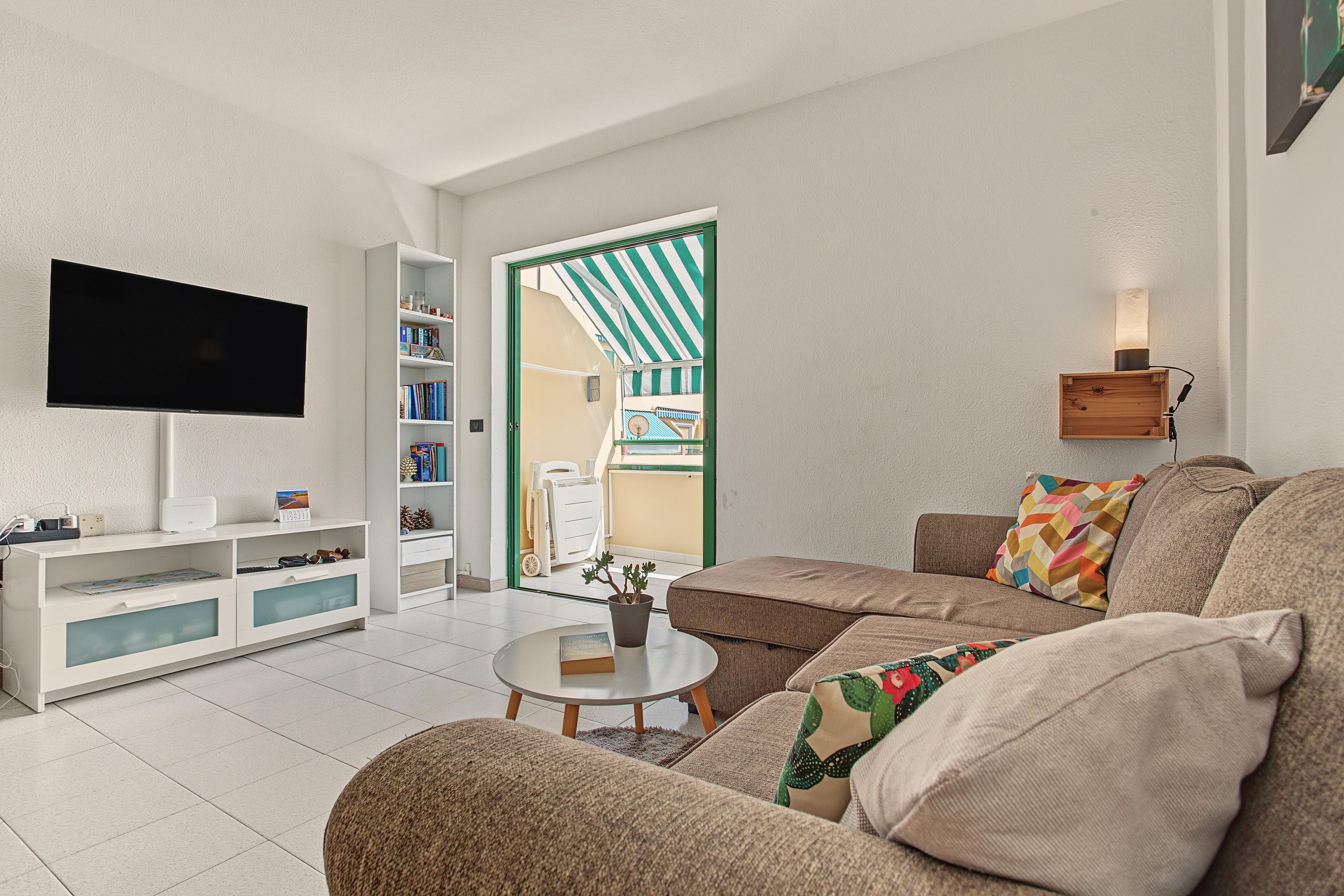 Ein modernes Wohnzimmer mit einem Flachbildfernseher, einer Bücherwand, einem Sofa mit gemusterten Kissen und einem kleinen Tisch mit einer Pflanze.