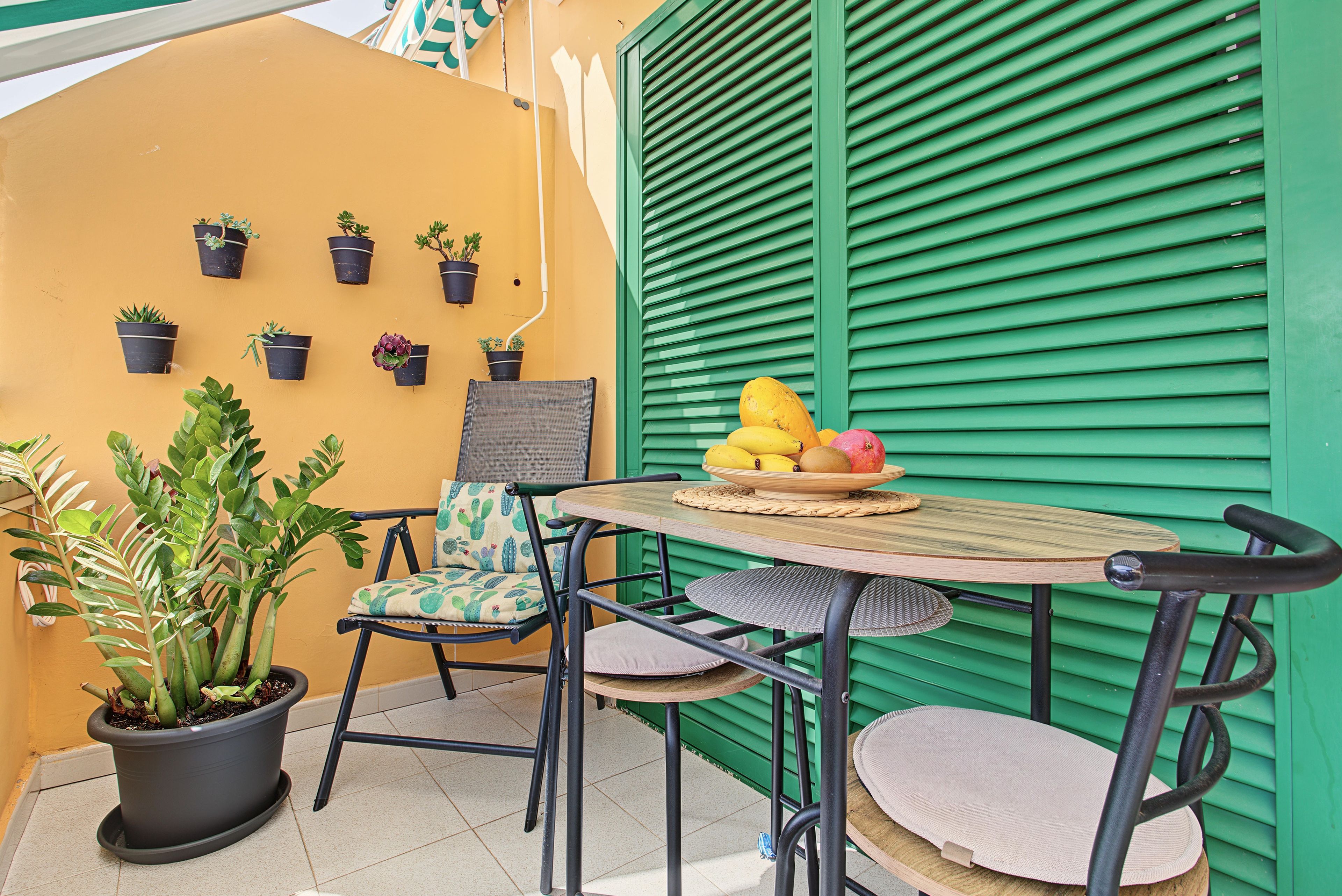 A small outdoor seating area with a table and chairs, surrounded by potted plants and a green shuttered wall.