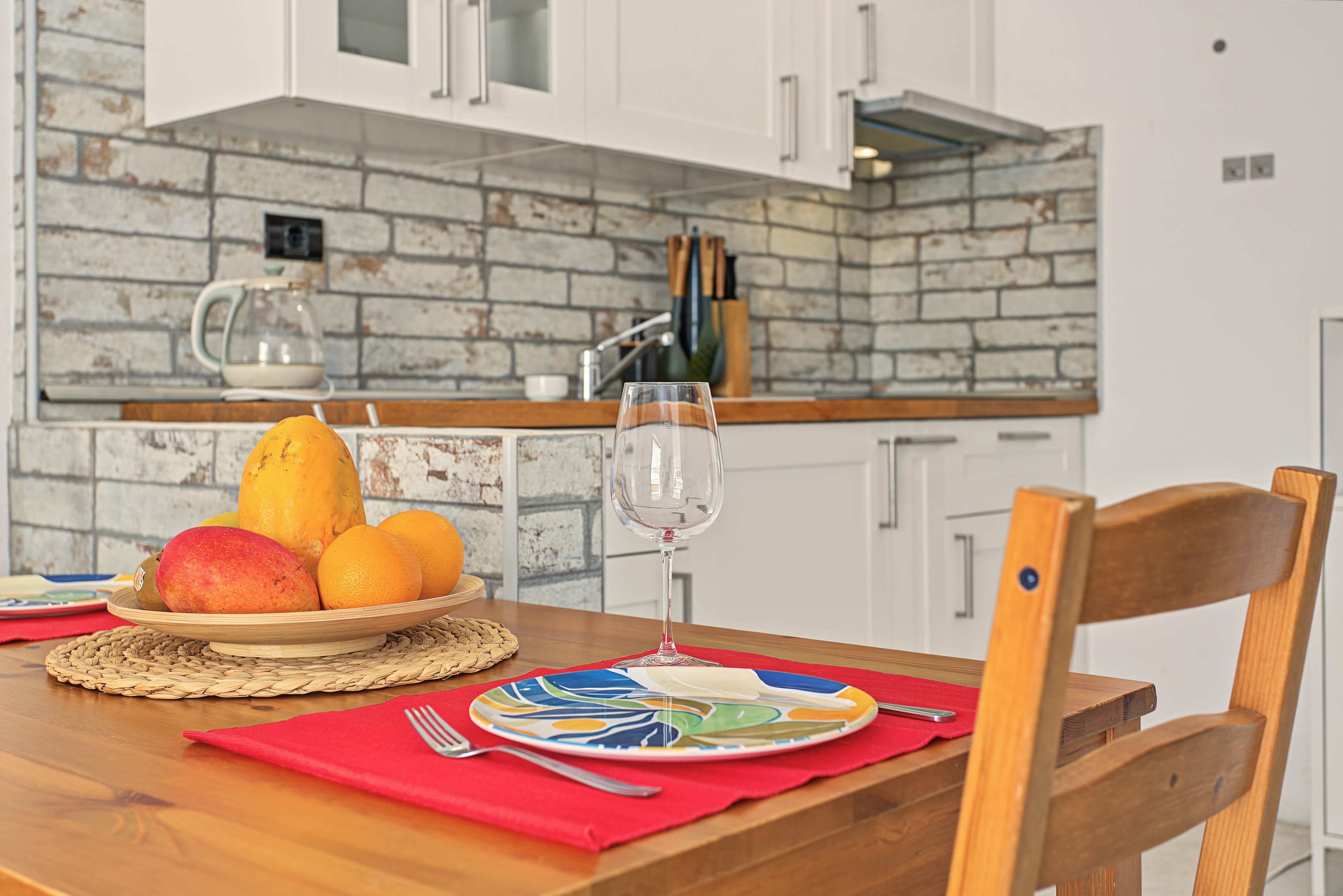 A kitchen with a brick backsplash, white cabinets, and a dining table set with fruit and a glass of water.