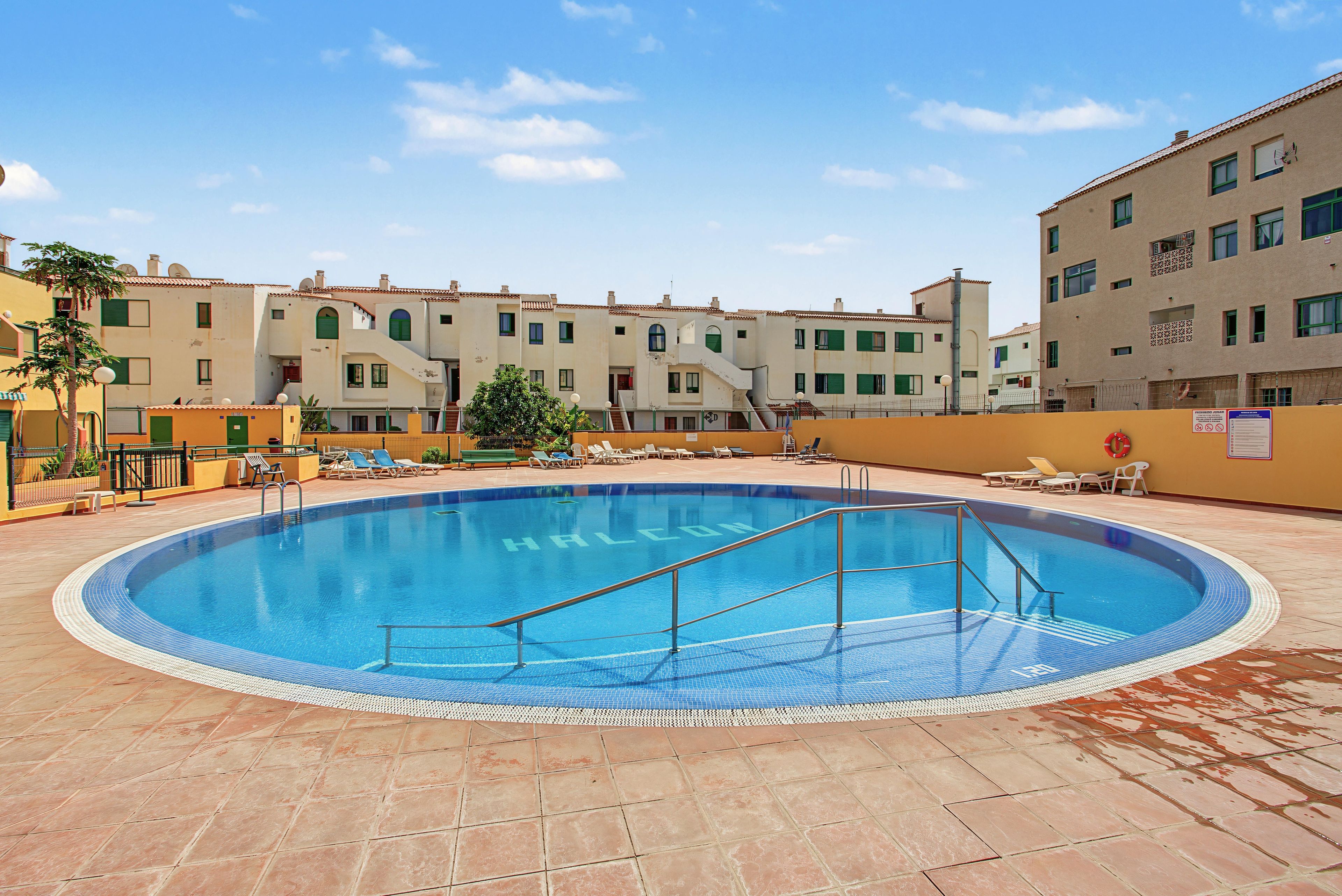 A swimming pool with a tiled edge, surrounded by lounge chairs and a building with multiple windows.