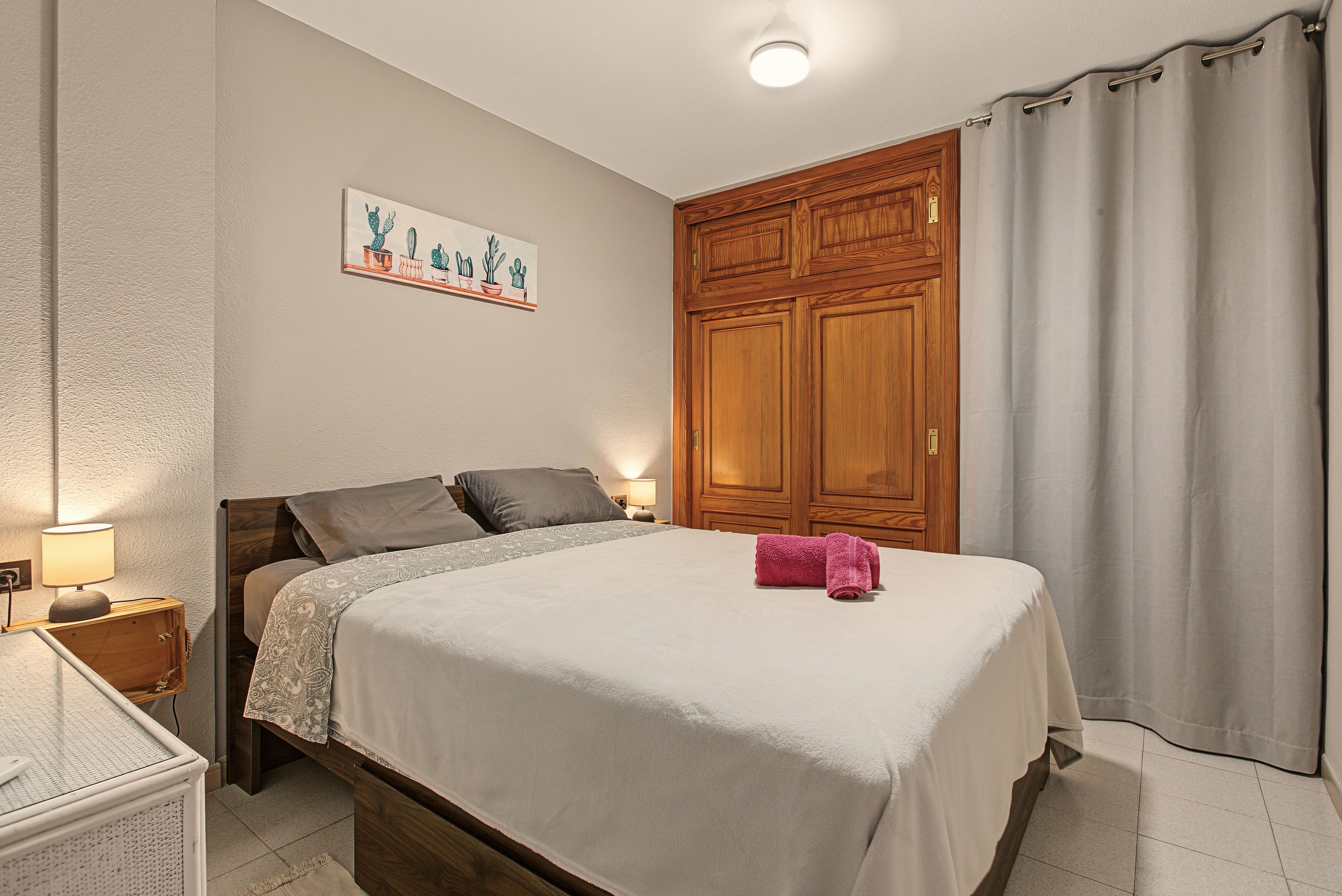Une chambre à coucher comprenant un lit, des tables de chevet, une commode, une lampe fixée au mur, une armoire en bois et une fenêtre avec des rideaux.