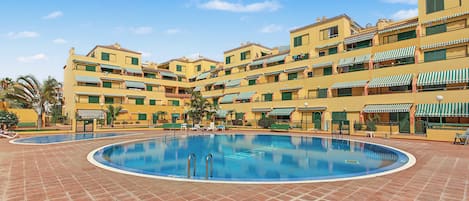 A hotel with a central swimming pool surrounded by tiled flooring, multiple balconies with green awnings, and a clear blue sky.