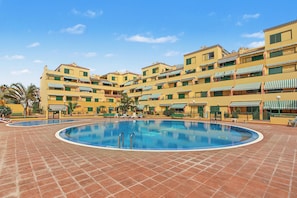 A hotel with a central swimming pool surrounded by tiled flooring, multiple balconies with green awnings, and a clear blue sky.
