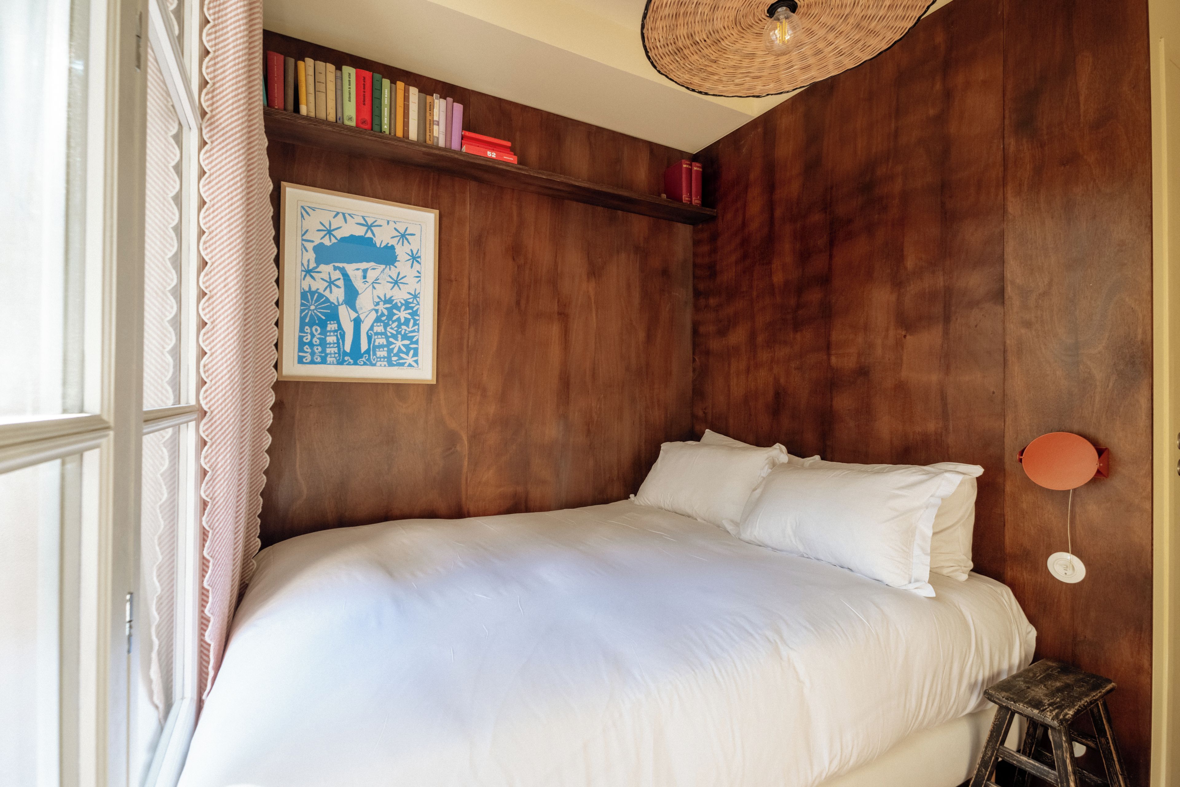 Une chambre à coucher avec un grand lit, un petit tabouret, une bibliothèque remplie de livres et un tableau encadré accroché au mur.