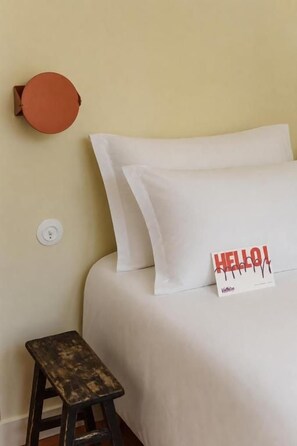 Ein Bett mit weißer Bettwäsche, ein kleiner Hocker und ein orangefarbenes Objekt an der Wand.