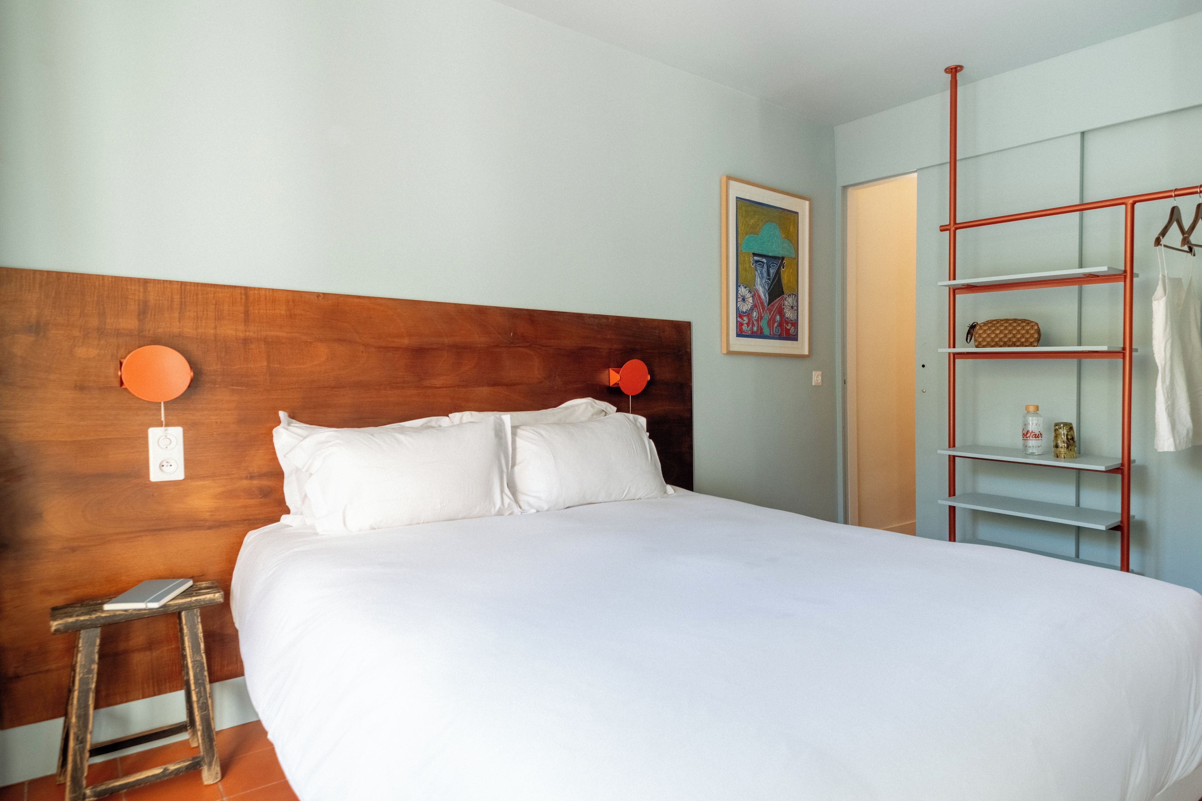 Habitació d'hotel amb un panell de fusta al capçal del llit, un llit amb llençols blancs, un petit tamboret, una prateleira amb articles de tocador i un quadre a la paret.