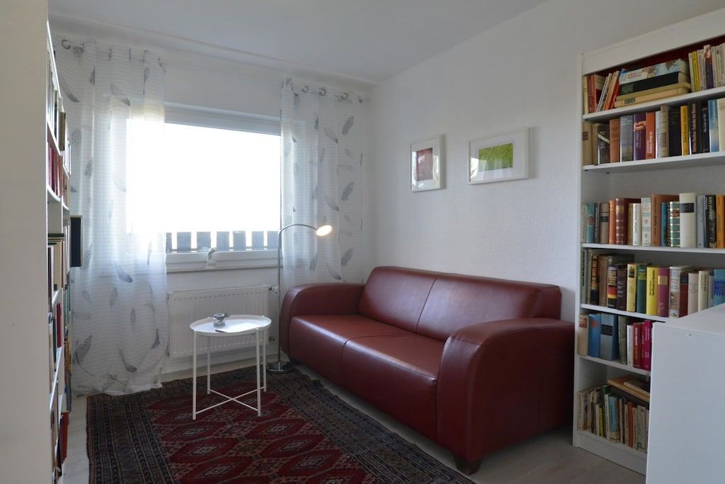 Una sala de estar con un sofá de cuero rojo, una mesita redonda pequeña, una estantería y una ventana con cortinas traslúcidas.