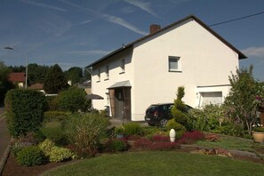 A two-story house with a car parked in the driveway, surrounded by a well-maintained garden with various plants and a lawn. - Holiday home in Eifel with 4 bedrooms (Dockweiler)