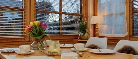 A dining table set with white plates, cups, and cutlery, a vase with flowers, and a lamp.