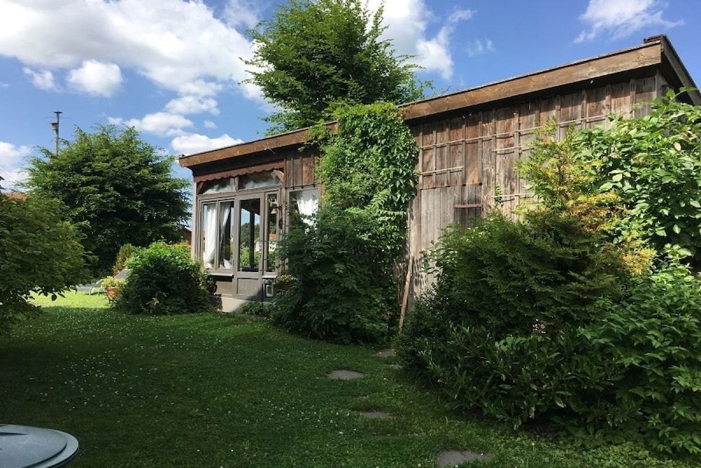 Un edificio rústico de madera con puerta de vidrio, rodeado de exuberante vegetación y un césped cuidado.