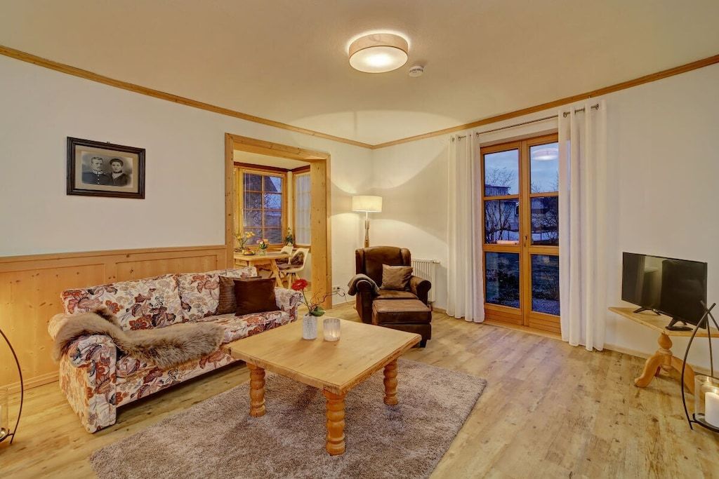 A cozy living room with a sofa, armchair, and coffee table. There is a television and a window with curtains.
