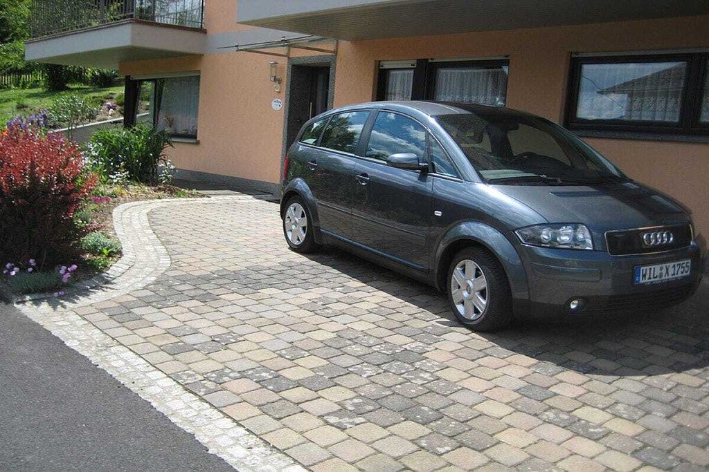 A compact car parked in front of a building with a paved driveway and a small garden area with flowers.