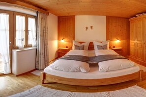 Ein rundes Bett aus Holz in einem Raum mit Holzwänden und einer geneigten Decke. Es gibt zwei Nachttische mit Lampen, ein Fenster mit Vorhängen und eine Heizung.