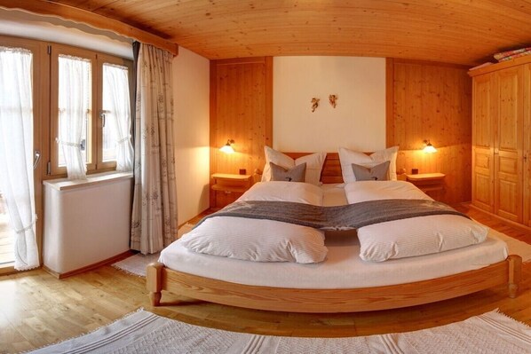 A circular wooden bed in a room with wooden walls and a sloped ceiling. There are two bedside tables with lamps, a window with curtains, and a radiator.