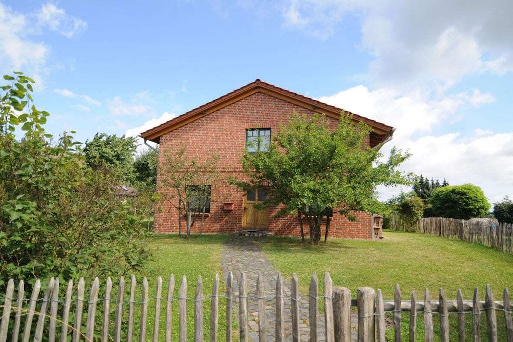 Un bâtiment en briques surmonté d’un toit à pignon, avec une clôture en bois et un chemin pavé menant à l’entrée.