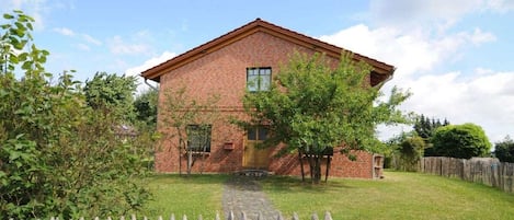 A brick building with a gabled roof, a wooden fence, and a paved pathway leading to the entrance.