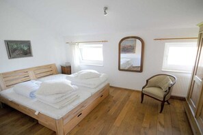 Ein Schlafzimmer mit einem hölzernen Bett, einem Sessel, einem Spiegel und zwei Fenstern.