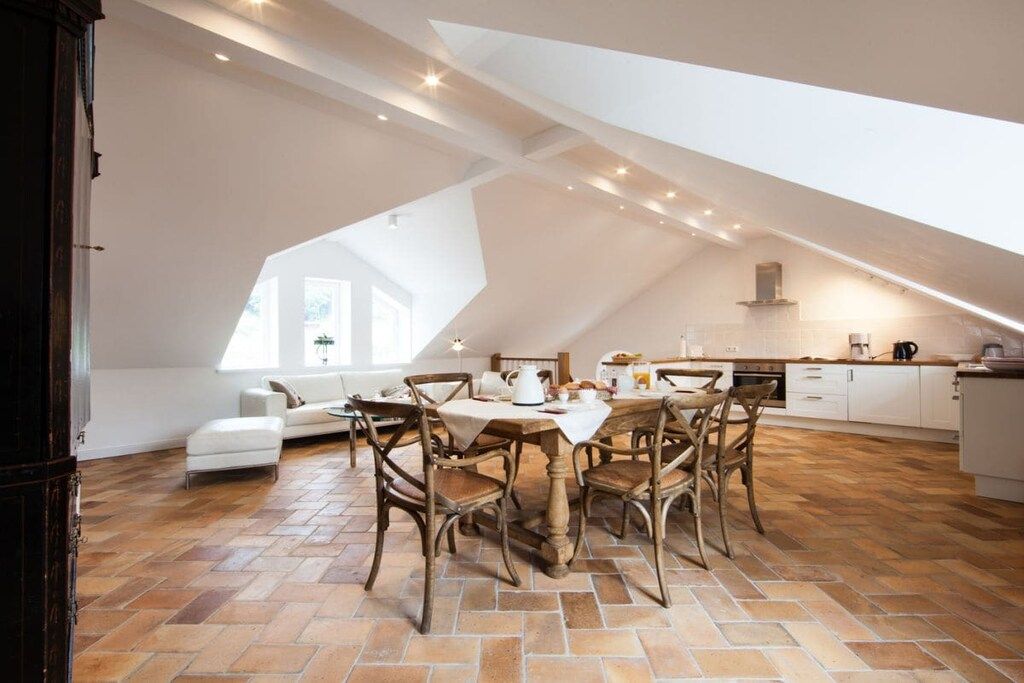 A spacious loft-style room with a high ceiling, a dining table with chairs, a sofa, and a kitchen area.