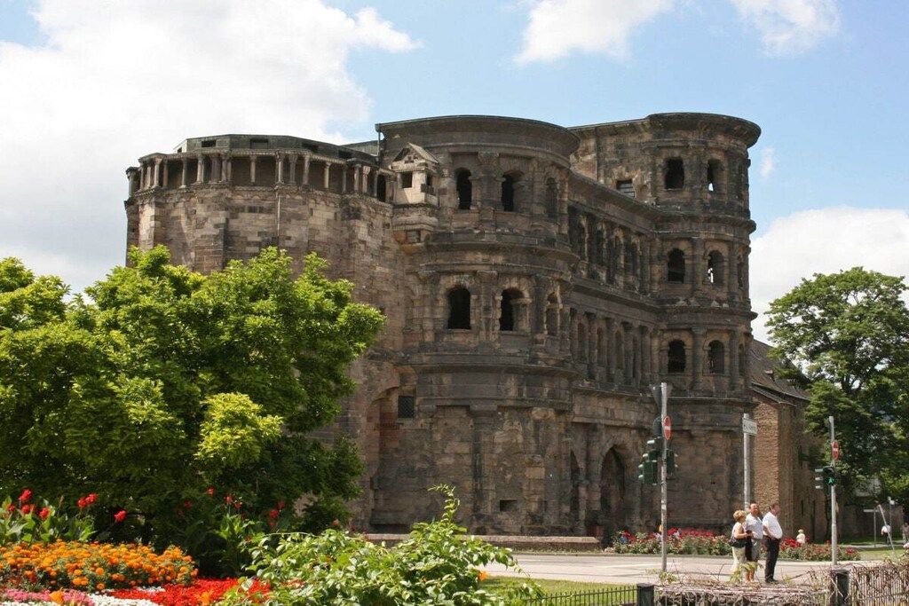 Un edificio histórico de piedra con múltiples arcos y una torre redonda, rodeado de abundante vegetación y plantas en flor.