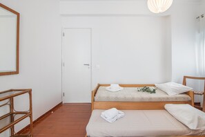 Ein schlicht eingerichteter Raum mit einem Bett, einem Holzstuhl, einem Korbstuhl und einer Tür.
