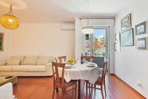 Ein Essbereich mit einem Tisch für zwei Personen, einer Couch im Hintergrund und Blick auf Gebäude durch ein Fenster.
