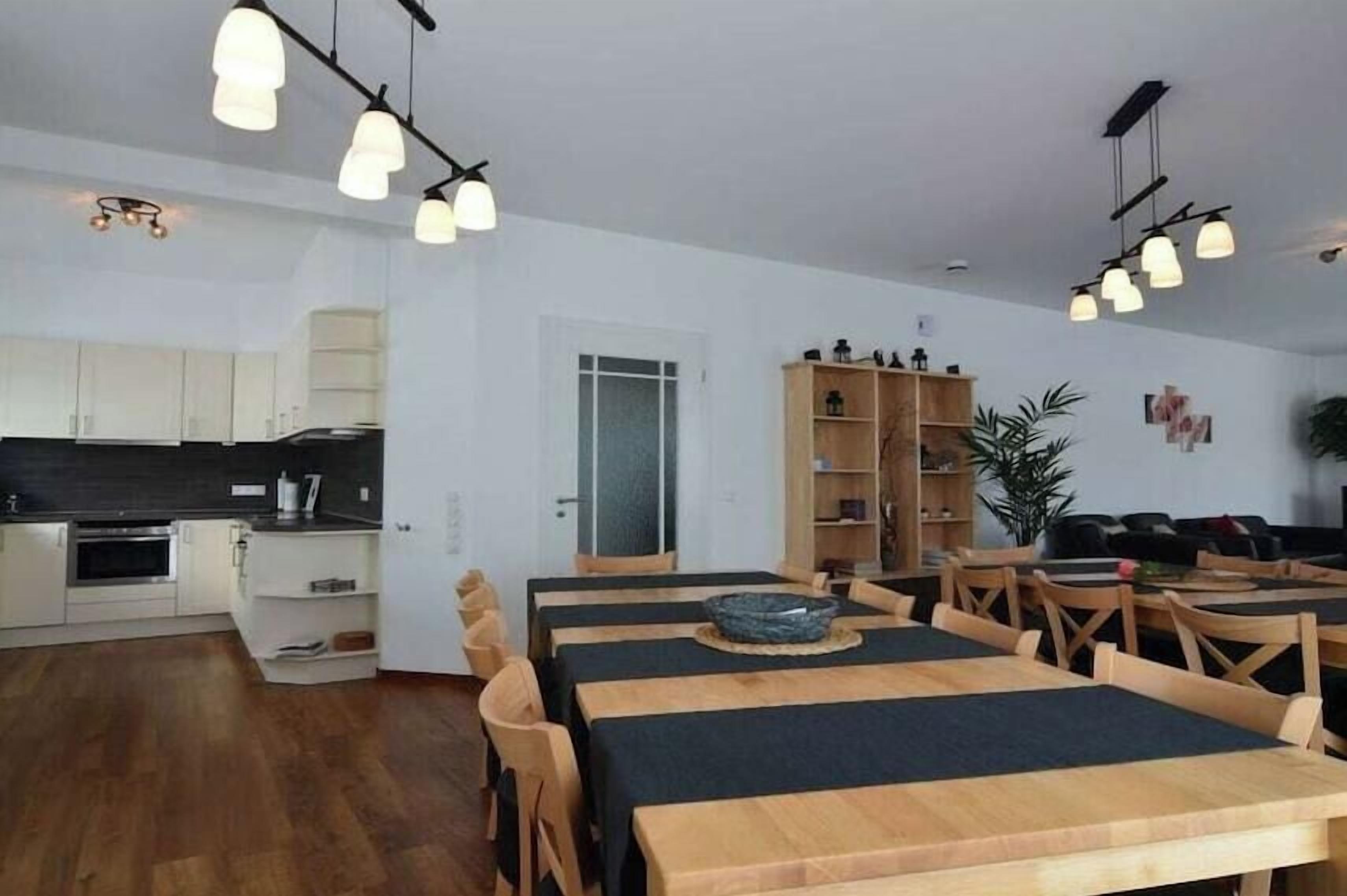 Et moderne køkken med hvide skabe og en sort bordplade, et spiseområde med træborde og stole, samt en reol med forskellige genstande.