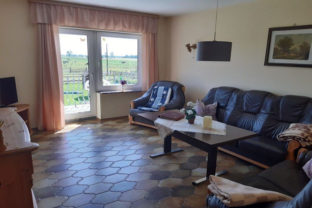 A living room with a sofa, a chair, a table with a vase, and a view of a farm through a glass door.