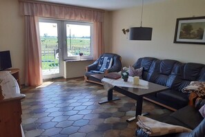 A living room with a sofa, a chair, a table with a vase, and a view of a farm through a glass door. - at the Wäcken holiday farm (Krummhörn-Grimersum)