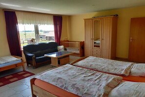 Ein Zimmer mit zwei Betten, einem Schlafsofa, einem Holzschrank, einem kleinen Tisch und Blick ins Freie.