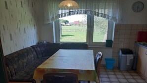 A dining area with a table and chairs, a sofa, a window with a view of the outdoors, a trash bin, and a clock on the wall. - at the Wäcken holiday farm (Krummhörn-Grimersum)