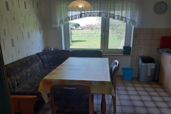 Ein Essbereich mit Tisch und Stühlen, einer Couch, einem Fenster mit Blick ins Freie, einer Mülltonne und einer Uhr an der Wand.