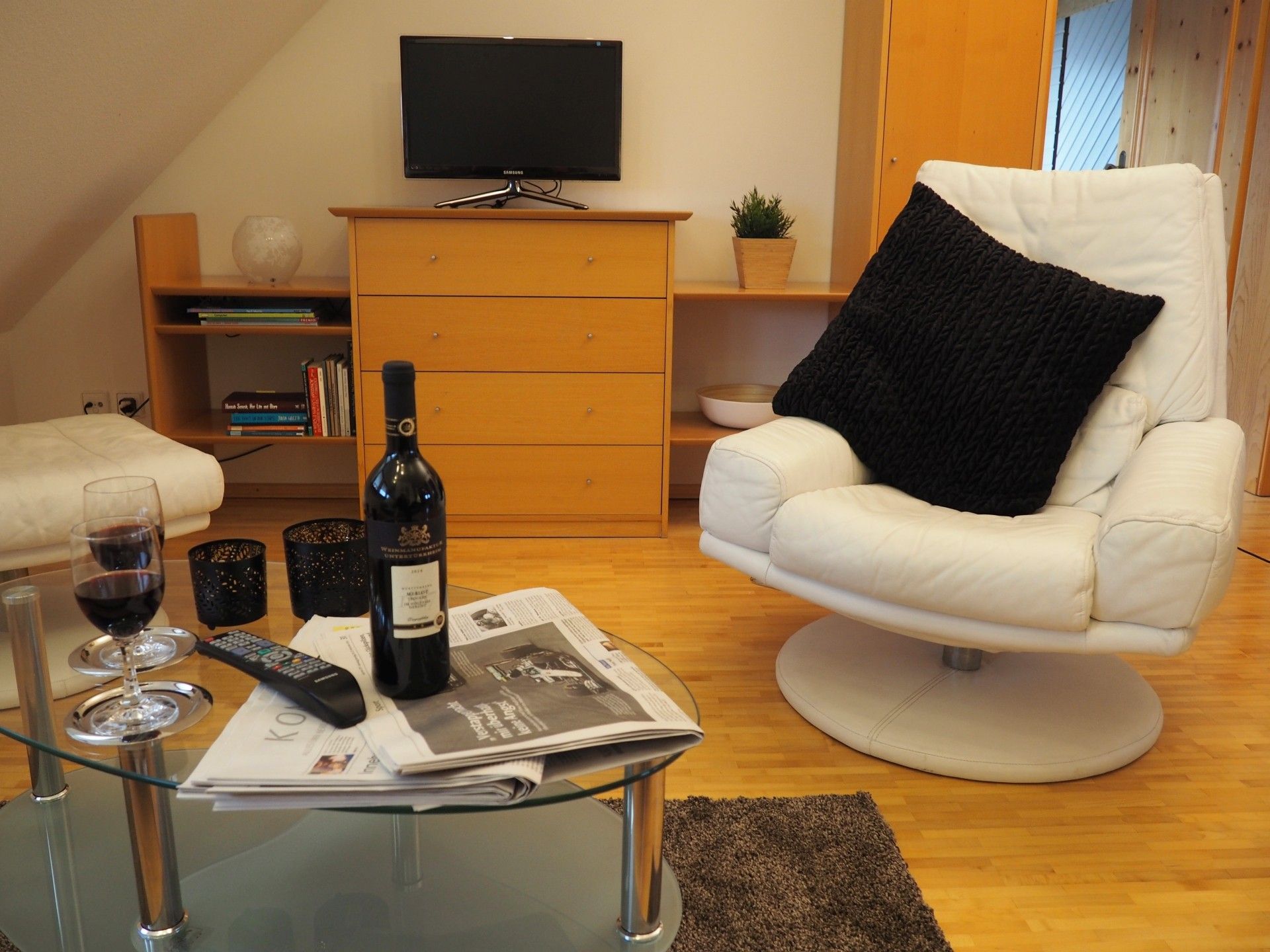Ein Wohnzimmer mit einem weißen Sessel, einem Glastisch mit einer Weinflasche, Gläsern und einer Fernbedienung.